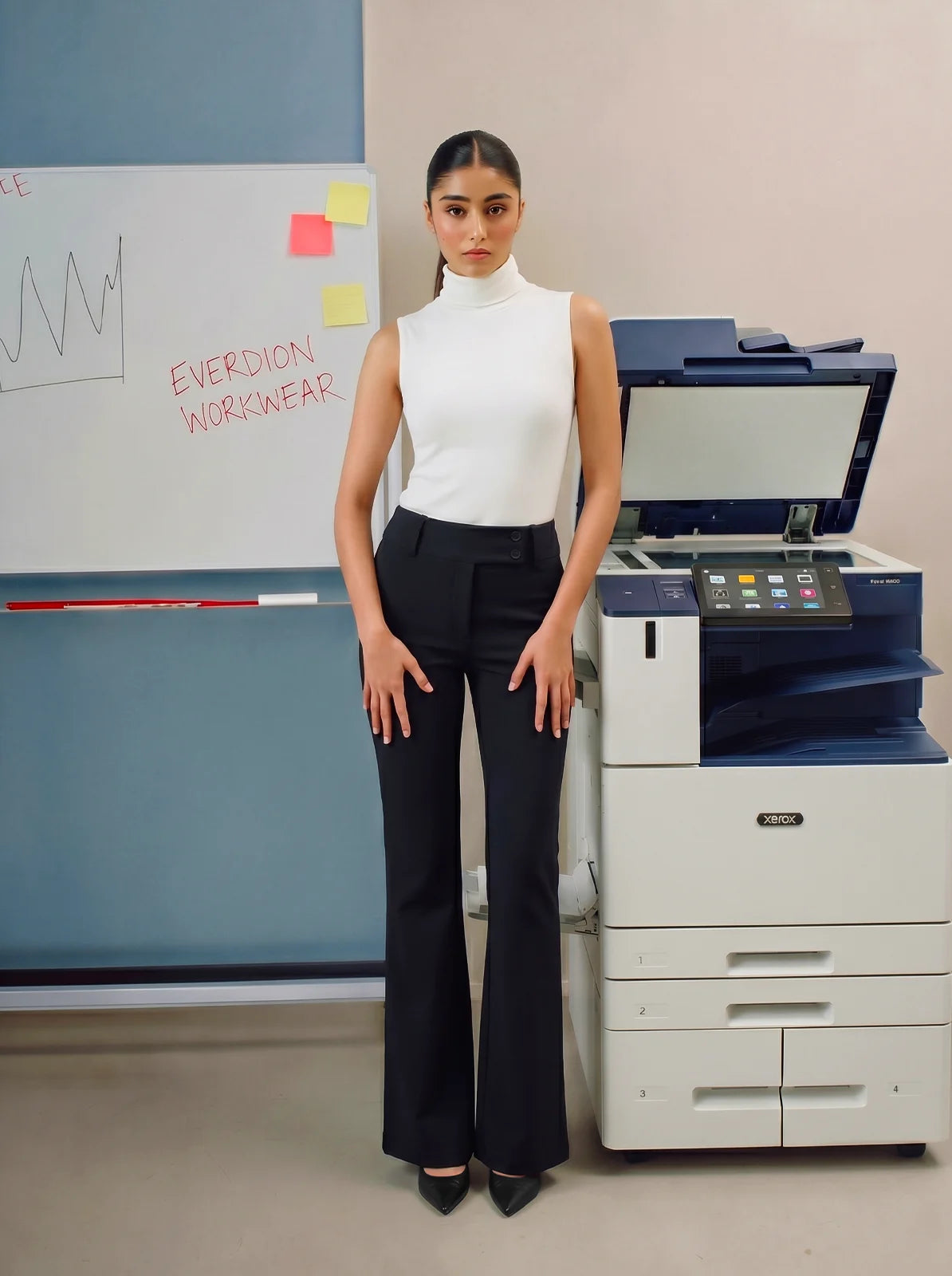 Woman in a white top and black pants standing next to a printer in an office setting.