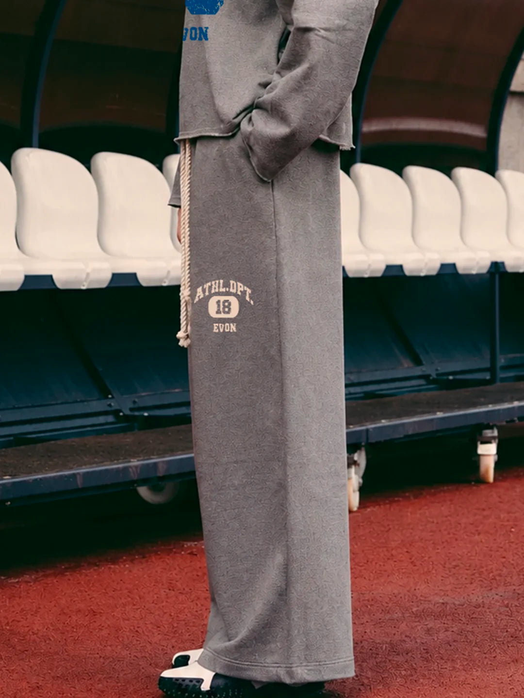 A person wearing a grey tshirt and matching wide-leg sweatpants in a stadium setting. The sweatpants feature a cream-colored drawstring and a cream graphic print on the left thigh that reads 'ATHLET.