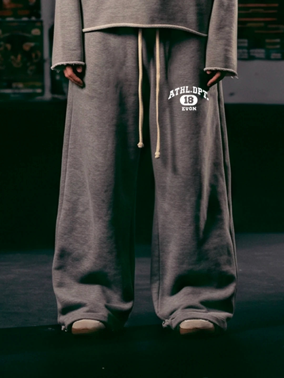 A person wearing loose grey sweatpants with a white 'ATHLETIC' graphic and drawstring, shown from the waist down in a dark indoor setting.