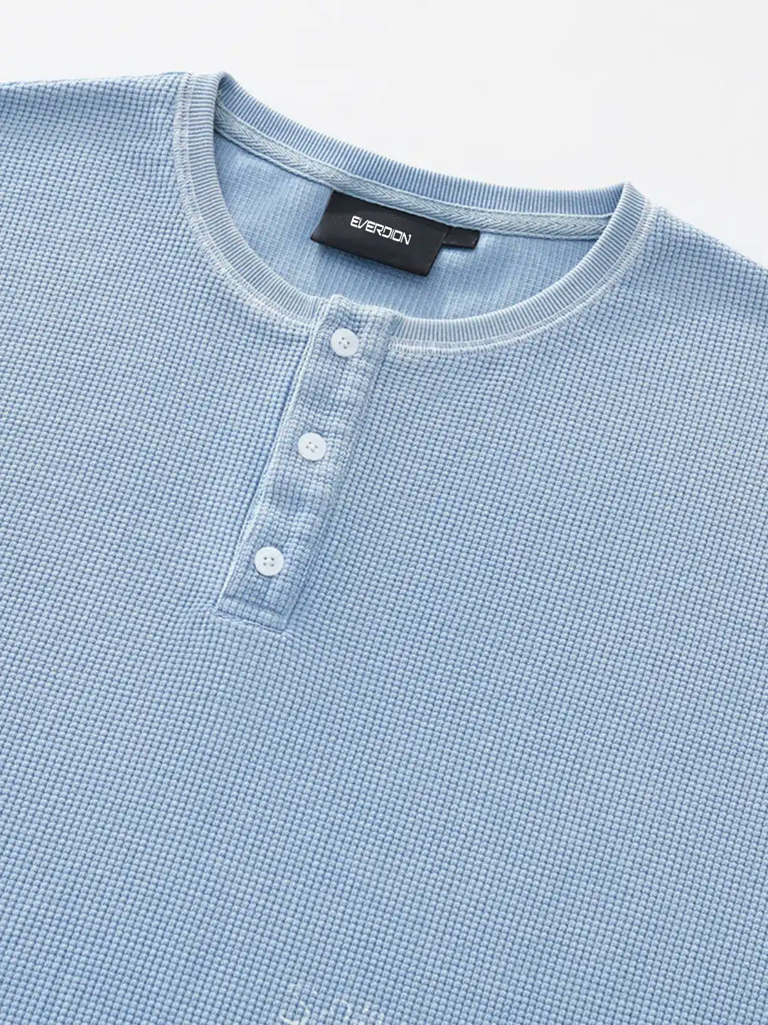 A close-up of a light blue waffle-knit henley t-shirt, featuring a three-button placket and a black brand label on the inner collar.