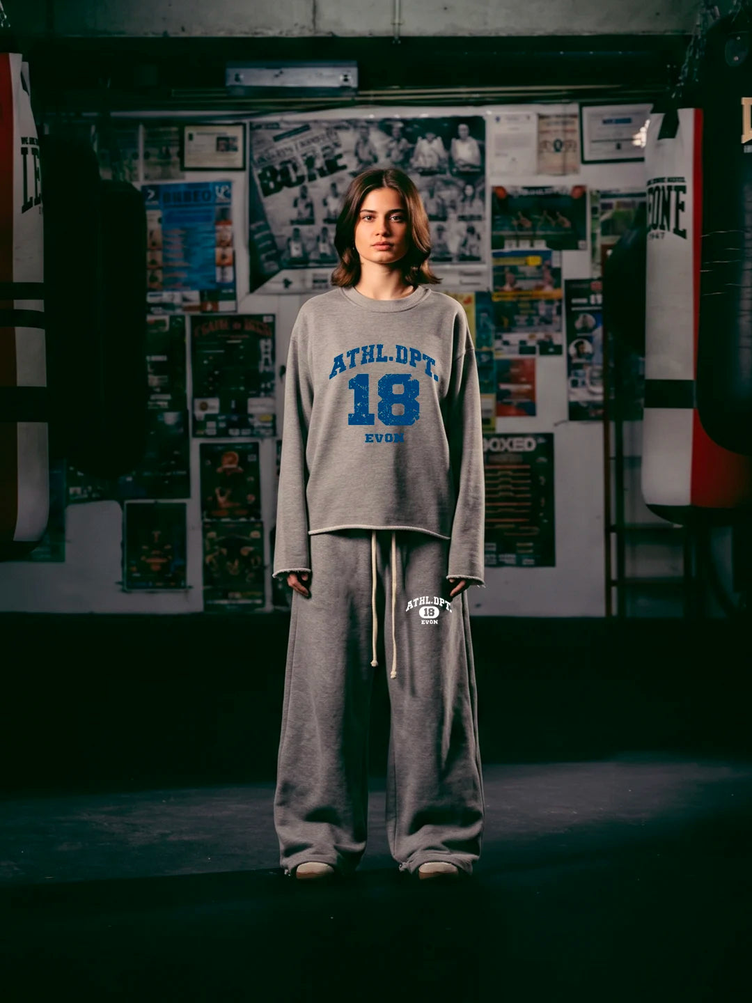 A women wearing a grey athletic tshirt and matching sweatpants. The tshirt features 'ATHLET 18 EVER' printed in blue on the front, and the sweatpants have a white logo on the thigh.