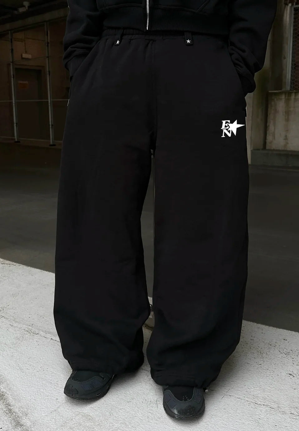 Black wide-leg sweatpants with a white embroidered logo on the left thigh, worn by a model in an urban setting.