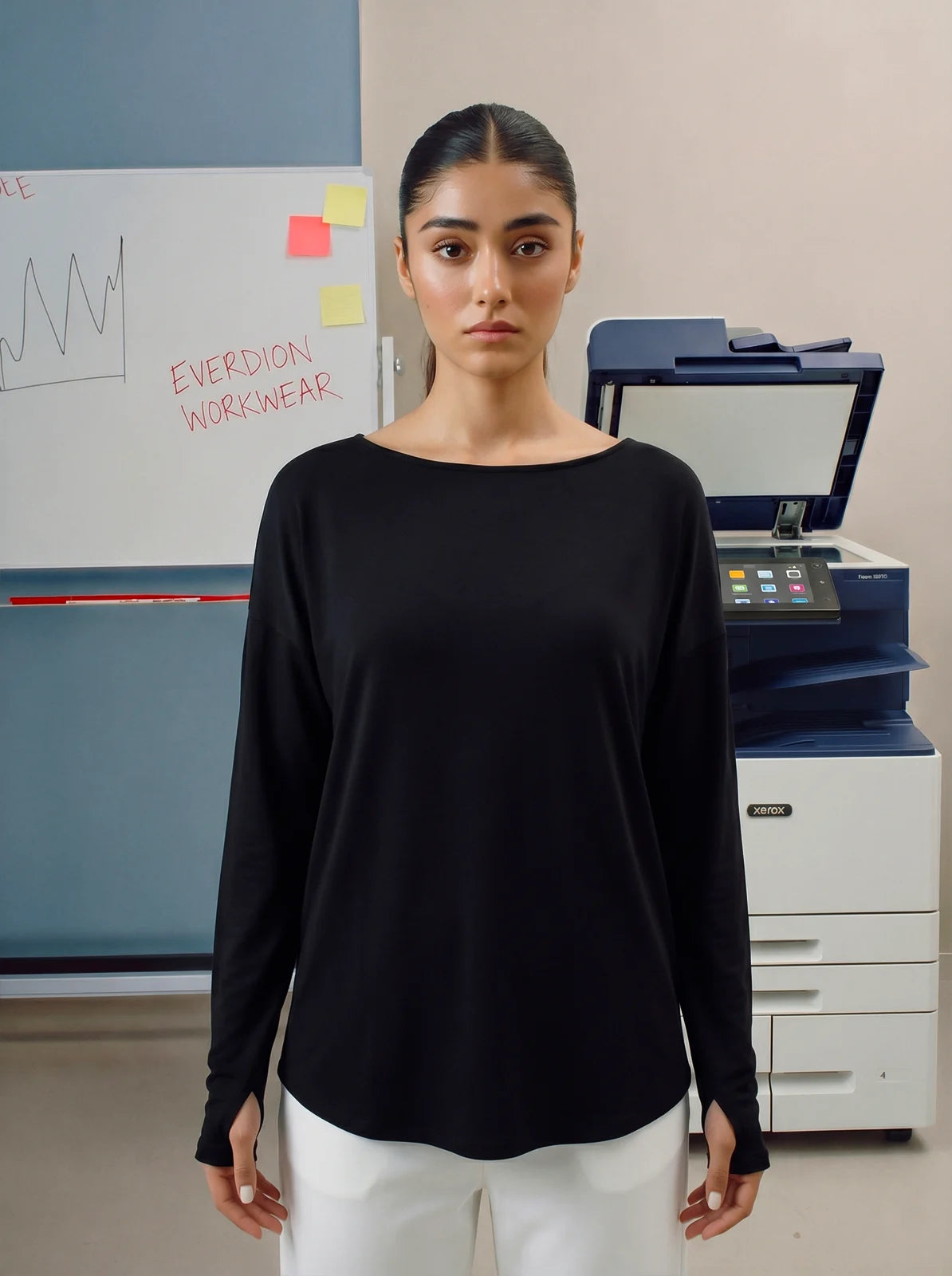Woman wearing a black long-sleeve top and white pants standing in an office setting.